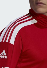 Red athletic jacket with a high neck, featuring white accents and three white stripes on the shoulders. Adidas logo in white on the sleeve.