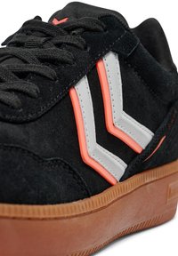 Svarta mockaskor med vita och neongula zigzag-accenter, med en strukturerad tan gummisula och platta svarta skosnören.