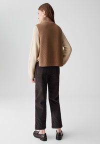 Brauner gerippter Strickwesten über einem cremefarbenen, lockeren Pullover, schwarze Cordhosen und dunkle Slipper; Modell von hinten vor einem hellen Hintergrund betrachtet.