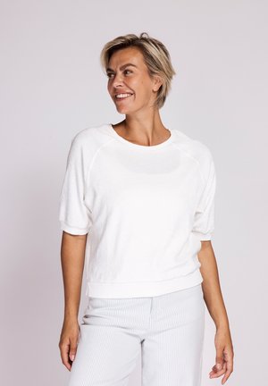 Smiling woman with short blond hair wearing a white short-sleeve top and light striped pants standing against a plain background.