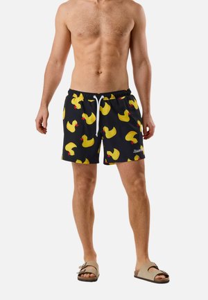 Skjorteløs mand iført sorte badebukser med gult gummidukke-mønster og beige sandaler, stående mod en ensfarvet baggrund.