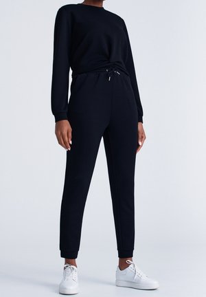Persoon draagt een zwarte top met lange mouwen en bijpassende zwarte joggingbroek met trekkoord in de taille, gecombineerd met witte sneakers tegen een effen achtergrond.