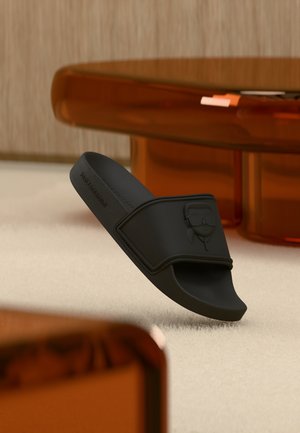 Crna slidna sandala s reljefnim dizajnom lica, naslonjena na svjetli tepih, s prozirnim narančastim stolom u pozadini.