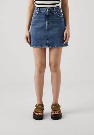 Femme portant une mini-jupe en jean bleu, un haut rayé et des sandales marron à boucle, debout devant un fond gris uni.