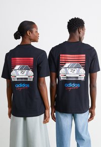 Melna T-kreka ar grafiku, kurā attēlots automobilis ar sarkaniem un zilajiem svītrām. Uzraksts ietver "adidas Originals" un skaitli "72".