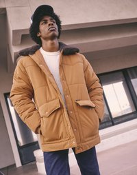 Bruine donsjas met een kraag gevoerd met bont, voorzien van grote zakken en drukknopen, gedragen over een grijze trui en blauwe spijkerbroek.