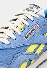 Μπλε σουέτ και συνθετικό αθλητικό παπούτσι με κίτρινες λεπτομέρειες. Διαθέτει σχέδιο με κορδόνια, υφαντή επιφάνεια και ετικέτα με το λογότυπο της Reebok.
