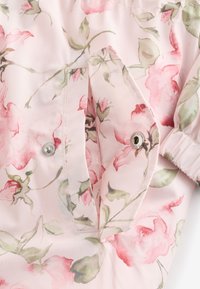 Imperméable fleuri rose présentant une texture lisse et brillante, une fermeture à pression, et un motif de roses avec des feuilles vertes. Détails cousus présents.