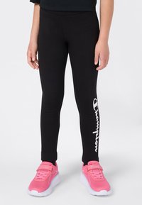 Kind in zwarte legging met het witte "Champion" logo op het linkerbeen en felroze sportschoenen met witte zolen.