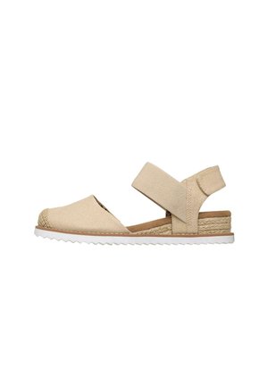 Sandalia plana beige con puntera cerrada, tira elástica en el tobillo, entresuela de yute tejida y suela de goma blanca con patrón estriado.