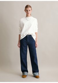 Junge Frau mit zurückgebundenem Haar, die ein weißes Langarmshirt, weite dunkelblaue Jeans, die am Boden umgeschlagen sind, und braune Schuhe trägt.