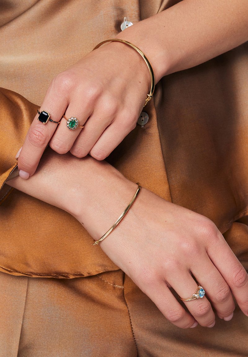 Złota metalowa bransoletka, dwa pierścionki: jeden z zielonym kamieniem otoczonym kamieniami, drugi z niebieskim kamieniem. Gładkie tekstury, kontrastujące kolory.