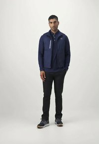 Sötétkék cipzáras dzseki sötétkék pólóing felett, fekete nadrággal és szürke sportcipővel. A dzsekin fehér cipzár részlet és logó található a mellrészen.