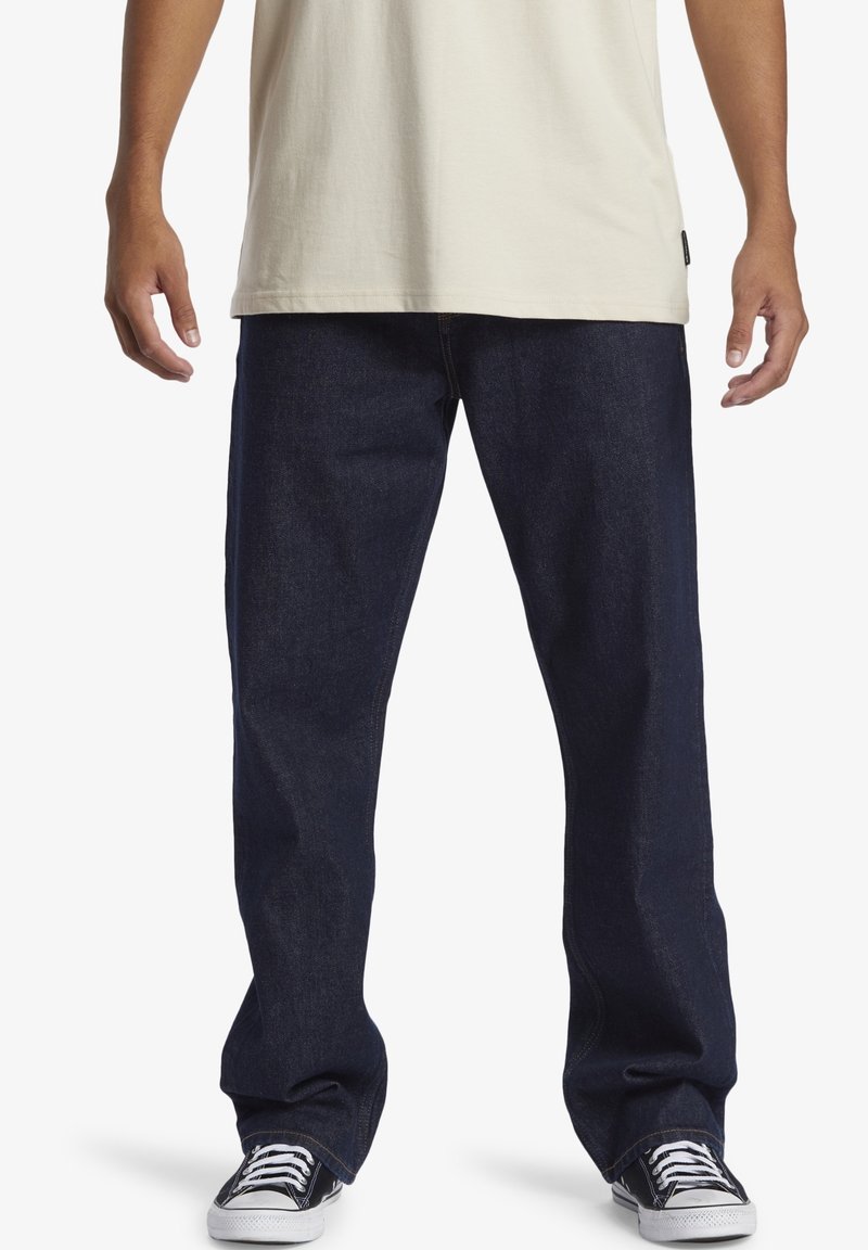 Homme portant un jean ample bleu foncé, une chemise beige et des baskets basses noires avec des lacets blancs, debout devant un fond blanc.