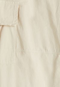 Tela de algodón beige con una superficie texturizada. Presenta un bolsillo con solapa y un detalle de costura cuadrada. Acabado suave y mate.