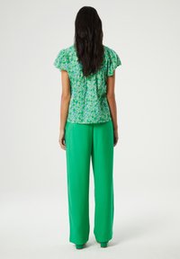 Groene bloemenblouse met korte ruchesleeves, gecombineerd met losvallende groene broeken. Gladde textuur, levendige kleuren en een ontspannen silhouet.