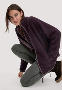 Paarse fleecetrui met een ontspannen pasvorm, contrasterende afwerking, ritszakken, en gecombineerd met groene leggings en bruine enkellaarsjes.