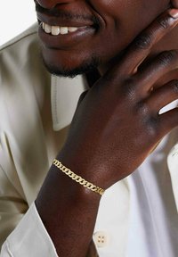 Personne souriante à la peau foncée portant une chemise beige et un bracelet en or, la main reposant doucement sur le côté de son visage.
