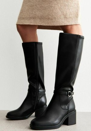 Botas de piel negras hasta la rodilla con tacón cuadrado, cremallera lateral y una correa con detalle de hebilla de metal. Textura suave con acentos mínimos.