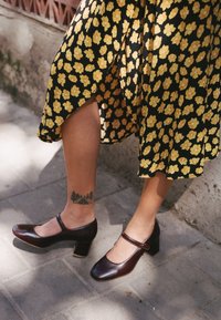 Hnědé kožené Mary Jane boty se čtveratou špičkou a blokovým podpatkem, spárované s černými šaty s žlutými květinovými vzory a viditelným tetováním na kotníku.