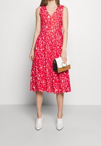Mujer vestida con un vestido rojo sin mangas con un patrón de salpicaduras blancas, botas blancas hasta el tobillo, sosteniendo un bolso blanco, marrón y negro.