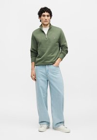 Mann trägt olivgrünes Sweatshirt mit Viertel-Reißverschluss, hellblaue Jeans mit weitem Bein, weiße Sneaker, steht mit einer Hand in der Tasche.