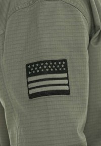 Tissu vert olive avec un motif texturé, featuring un patch brodé noir du drapeau américain avec des rayures et des étoiles blanches.