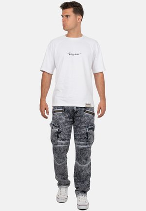 T-shirt blanche à manches courtes avec logo, associée à un pantalon cargo gris comportant de multiples poches et des détails zippés, portée avec des baskets blanches.