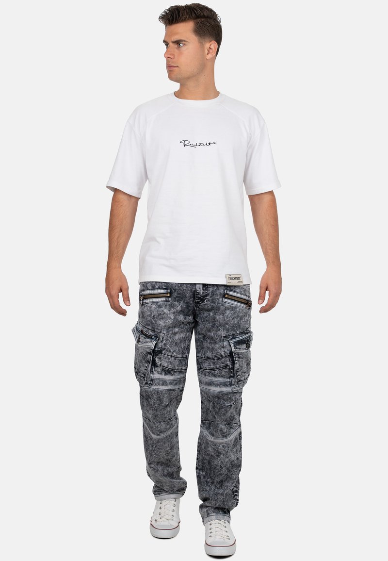 T-shirt bianco a maniche corte con logo, abbinato a pantaloni cargo grigi con molteplici tasche e dettagli con zip, indossato con sneaker bianche.