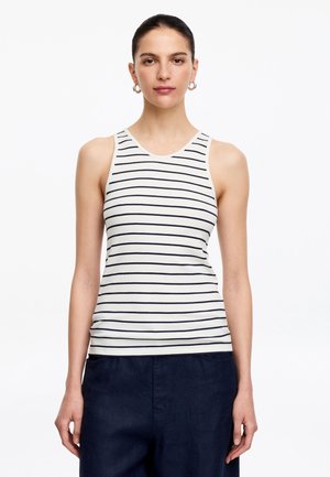 Femme aux cheveux foncés portant un haut sans manches blanc avec des rayures horizontales noires et un pantalon foncé, debout devant un fond blanc uni.