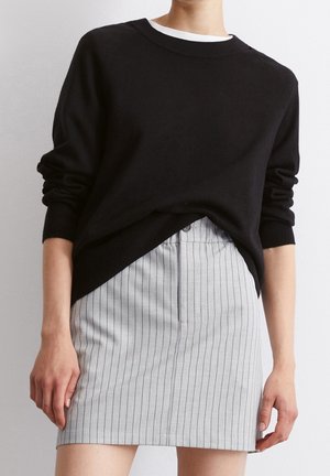 Persoon draagt een zwarte trui met lange mouwen over een wit hemd en een hooggetailleerde grijze rok met verticale zwarte strepen.