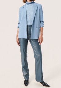 Blazer azul claro con solapas de pico y dos bolsillos frontales, combinado con un suéter de cuello alto azul claro y pantalones ajustados de imitación piel en tono teal.