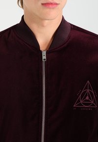 Giacca bomber bordeaux con una texture liscia, colletto a coste e zip anteriore argentata. Presenta un logo geometrico di colore rosso scuro.
