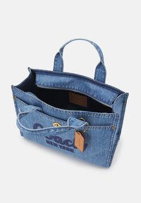 Sac cabas en denim bleu avec deux poignées, poche avant avec fermeture pression, compartiment principal zippé et une étiquette de marque en cuir suspendue.