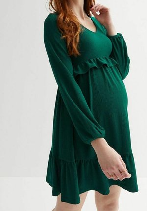 Pregnant woman wearing a long-sleeve, dark green, knee-length dress with ruffle details at waist and hem.