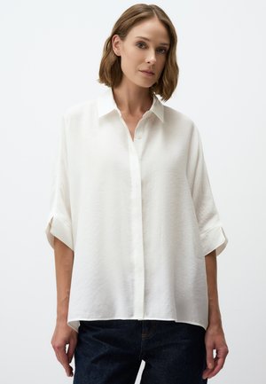 White button-up shirt with a loose fit, short rolled sleeves, and a textured fabric. Paired with dark denim jeans.