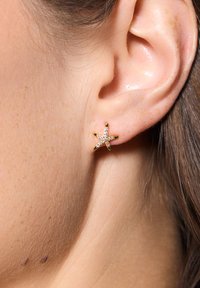 Złota kolczyk w kształcie rozgwiazdy ozdobiony małymi, przezroczystymi cyrkoniami, umieszczony na zbliżeniu ludzkiego ucha. Gładka tekstura skóry.