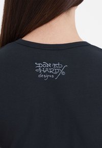 Musta puuvilla t-paita rentoon istuvuuteen, jossa on tekstuurilla painettu valkoinen logo "DON ED HARDY designs" selkäkaulan kohdalla.