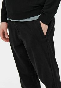 Zwarte corduroy broek met een relaxte pasvorm, voorzien van zijzakken en een gestructureerd oppervlak. Gedragen met een grijs, gelaagd shirt eronder.