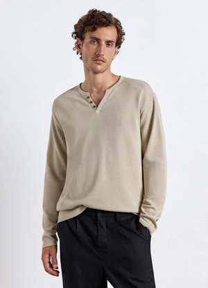 Uomo con capelli ricci indossa una camicia beige a maniche lunghe con bottoni e pantaloni neri, in piedi con una mano in tasca su sfondo bianco.