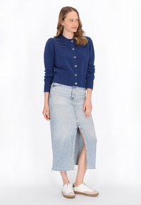 Cardigan lavorato a maglia blu navy con bottoni a forma di cuore, abbinato a una gonna di jeans azzurri chiari con spacco frontale e orlo sfilacciato. Sneakers bianche.