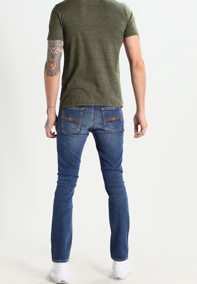 Homme debout pieds nus, face à dos, portant un t-shirt vert, un jean bleu avec poches arrière et un tatouage sur l'avant-bras gauche.