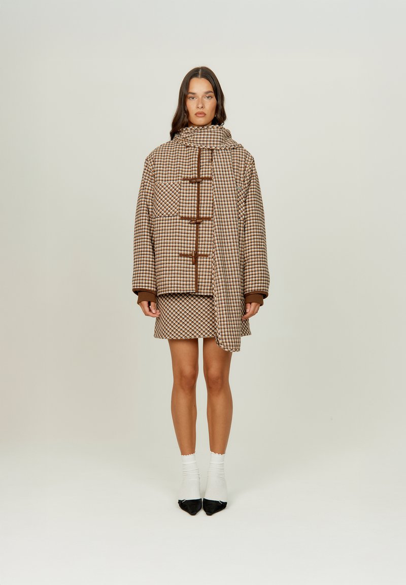 Καφέ καρό παλτό με μεγάλα κουμπιά, φορεμένο πάνω από φούστα σε ταιριαστό μοτίβο. Διαθέτει ψηλό γιακά, φαρδιά μανίκια και απαλή υφή.