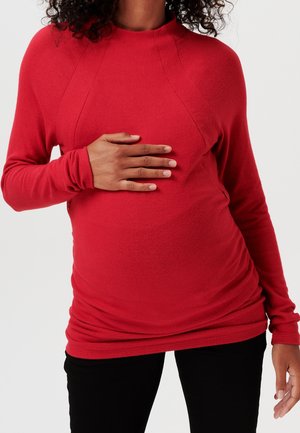 Punainen pitkähihaisen paita, joka on valmistettu pehmeästä kankaasta, pyöreällä pääntiellä, sivuilla rypytyksiä ja istuvat hihat. Paita on päällä raskaana olevan vatsan päällä.