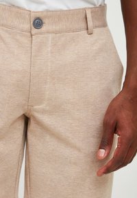 Pantalons beiges à motifs avec un design à carreaux texturé, fermeture à bouton et poches latérales, avec une finition en tissu lisse.