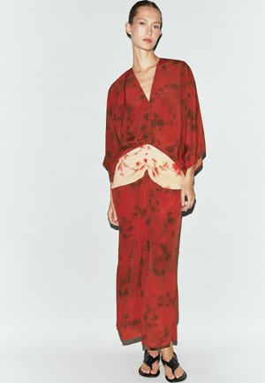 Frau steht und trägt ein rotes Batik-Maxikleid mit cremefarbenem, gerafftem Detail an der Taille und schwarzen gewebten Sandalen vor einem schlichten hellen Hintergrund.
