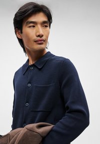 Ναυτικό πλεκτό ζακέτα με γιακά, περιλαμβάνει τέσσερα κουμπιά, τσέπη στο στήθος και υφή υφάσματος. Το άτομο κρατάει ένα καφέ παλτό.