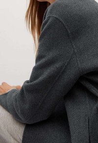 Personne aux longs cheveux portant un manteau en laine gris foncé, assise les mains posées sur ses genoux devant un fond clair uni.