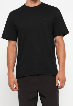 Homme portant un t-shirt noir uni à manches courtes avec un petit logo discret sur la poitrine gauche et un pantalon foncé sur fond blanc.