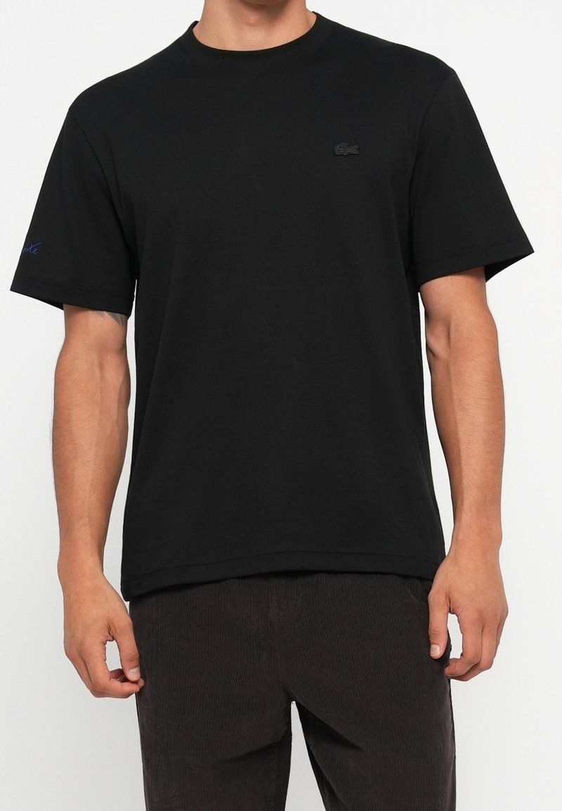 Homme portant un t-shirt noir uni à manches courtes avec un petit logo discret sur la poitrine gauche et un pantalon foncé sur fond blanc.